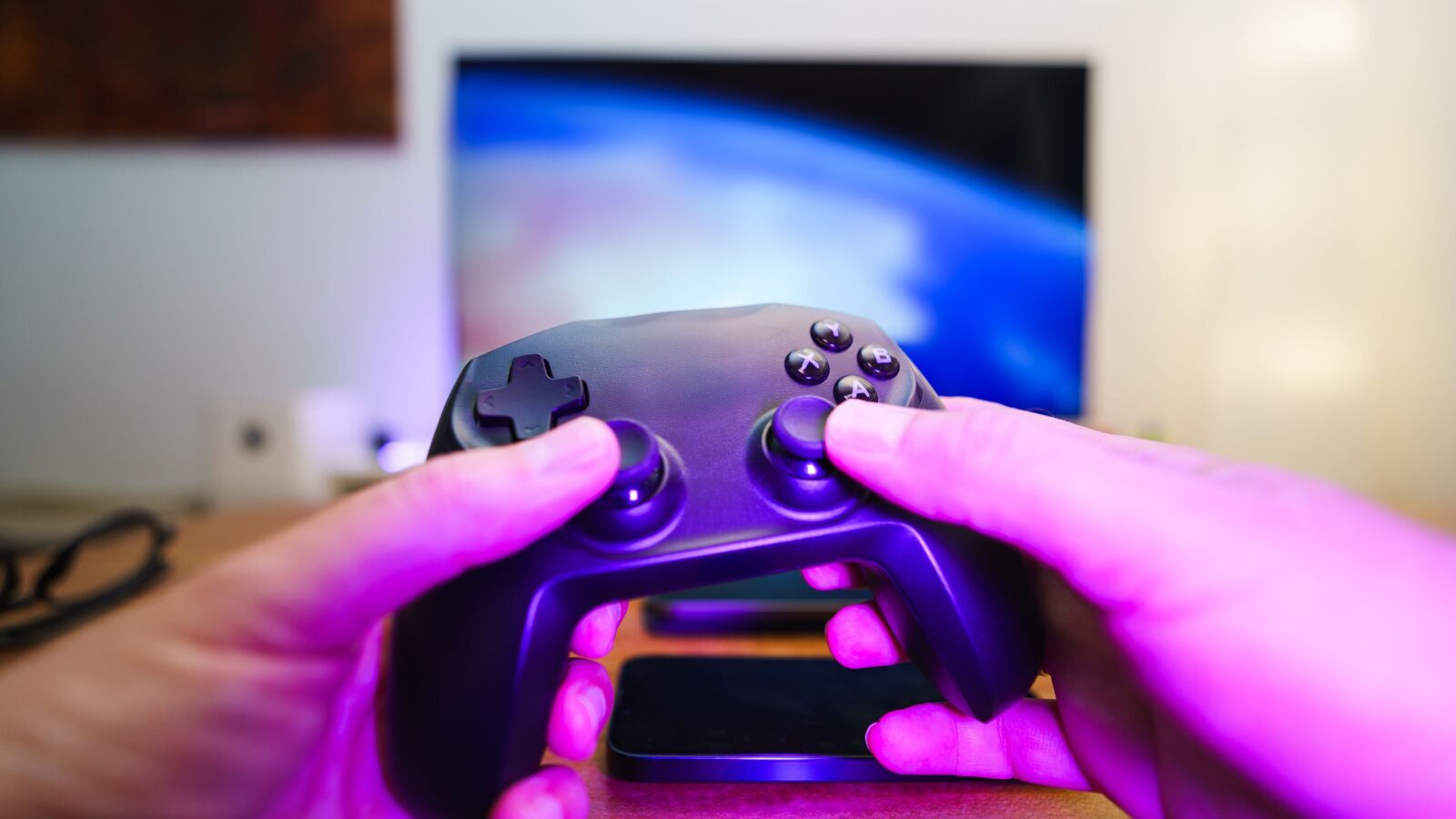 Hands holding a console controller in front of a lit TV, with purple night-time ambient light