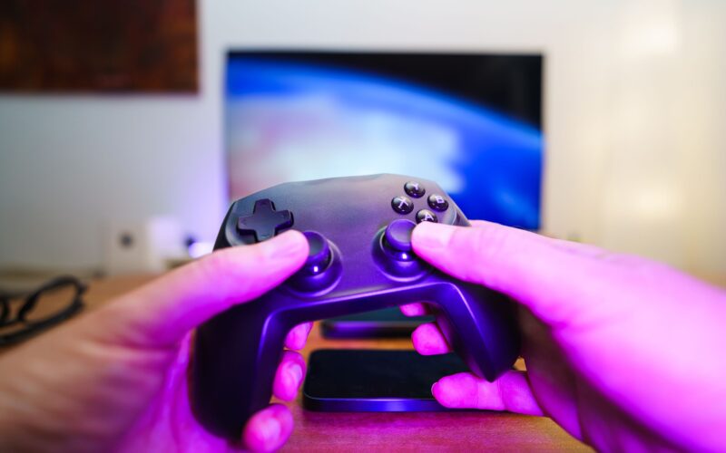 Hands holding a console controller in front of a lit TV, with purple night-time ambient light