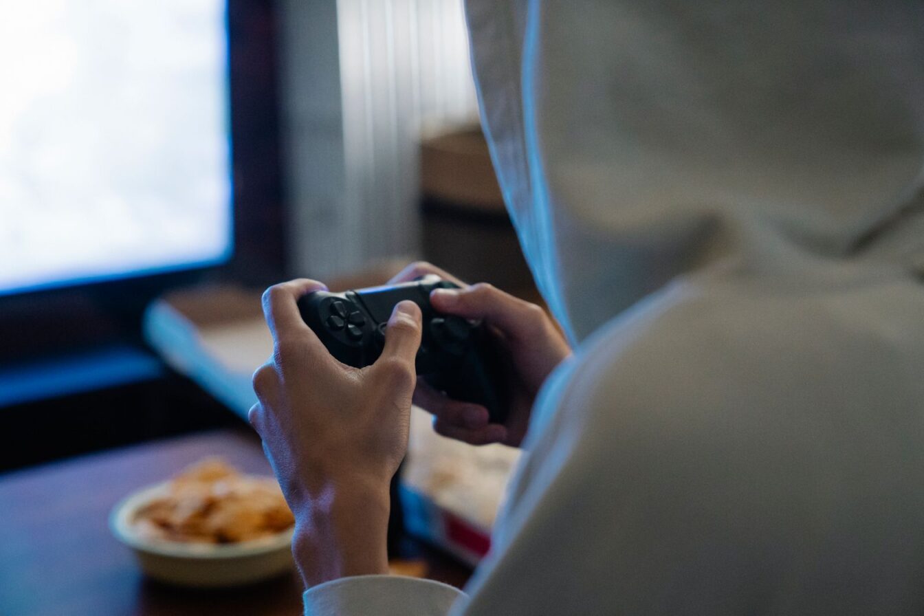 Hooded person seen from behind, playing at night in a dark room with a controller in hand