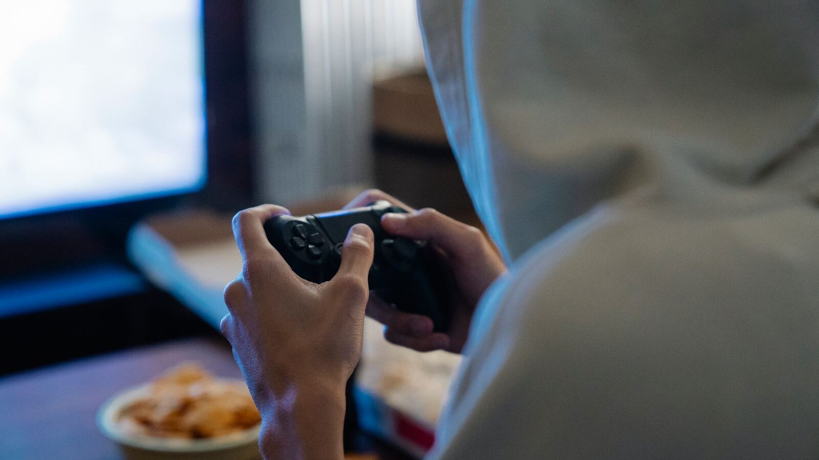 Hooded person seen from behind, playing at night in a dark room with a controller in hand
