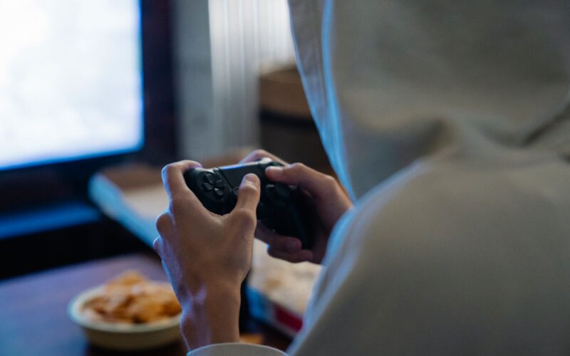 Hooded person seen from behind, playing at night in a dark room with a controller in hand
